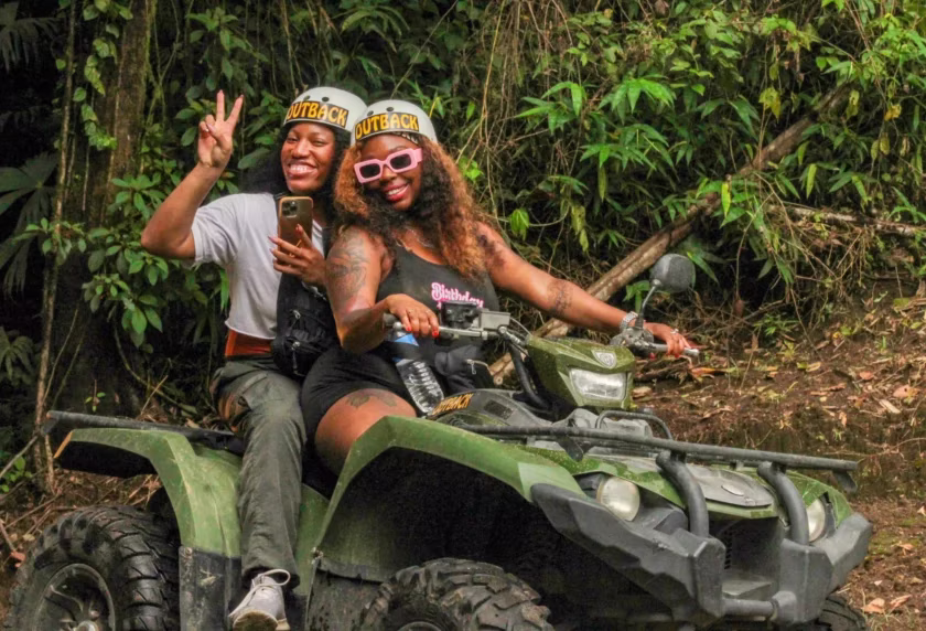 Happy female travelers riding an ATV and waving at the camera in a farm setting in La Fortuna, Costa Rica