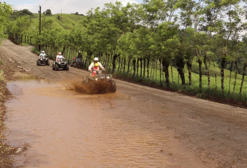 Father and son riding an ATV through a muddy puddle with more riders behind on a gravel road in Monterrey, La Fortuna, Costa Rica