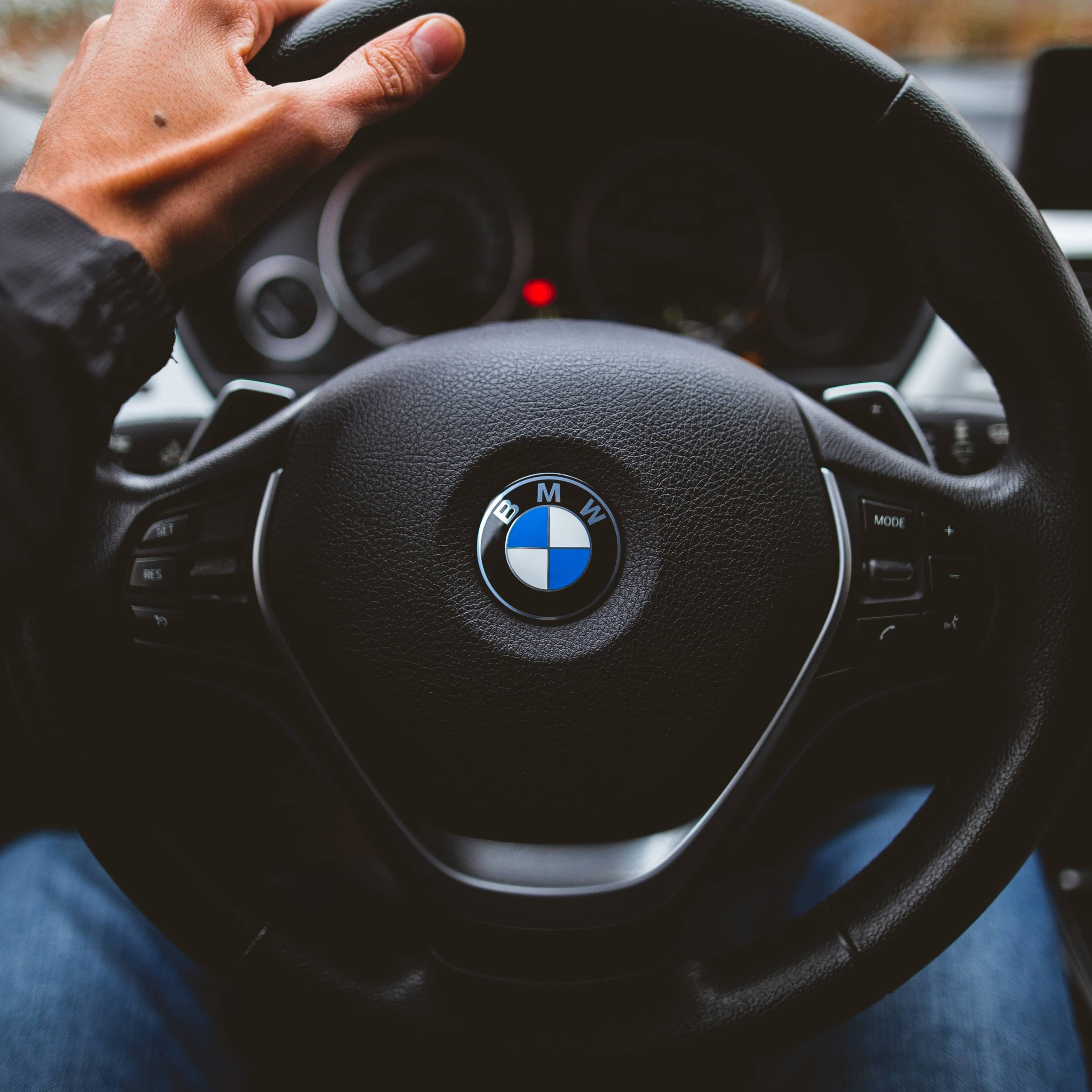 Steering wheel with BMW logo. A driver's left hand is on the wheel.