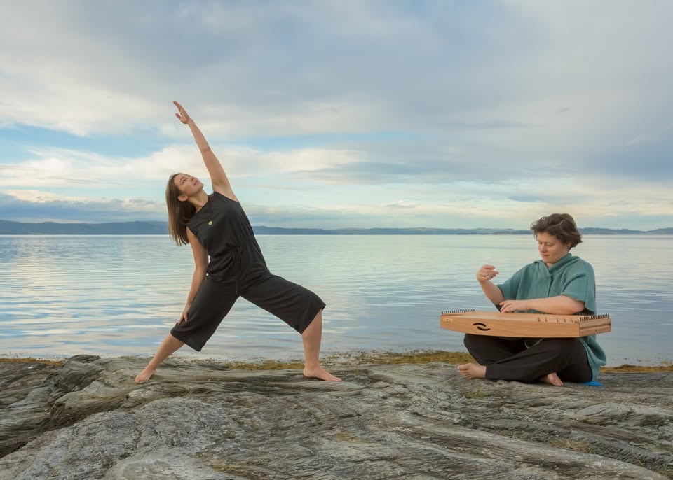 A person doing a yoga pose and another person playing Monochord in front of the fjord