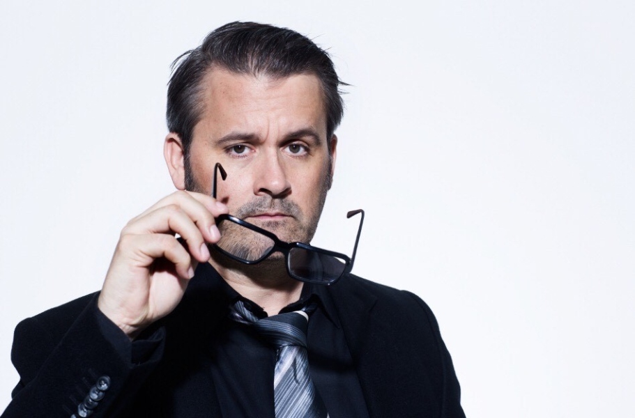 Portrett av hvit ung mann, mørkt hår, stubbskjegg, med briller i hånda