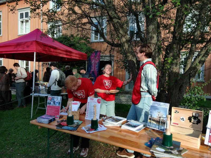 Foto viser stand med materiale fra Fortidsminneforeningen og frivillige fra Kulturnatt 2025. I bakgrunn av bilde ser man besøkende som ser på bøkene tilhørende antikvarisk bokloppis. 