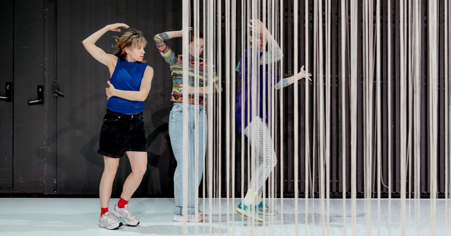 Tre personer danser. På høyre side av bildet henger et "spagetti"-forheng og dekker to av personene.