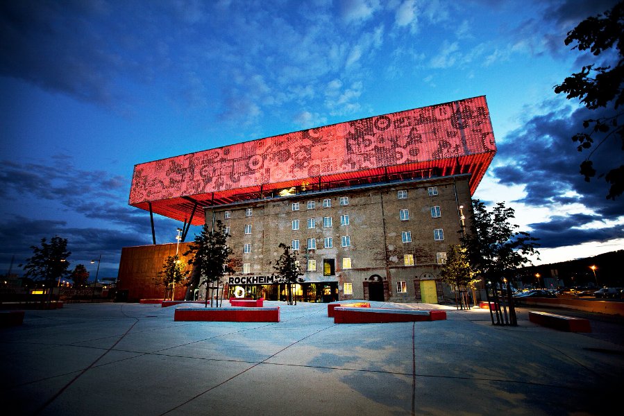 Bilde av Rockheim i den blå timen. Taket har rød dekor, det er trær foran bygget og skyer på himmelen.