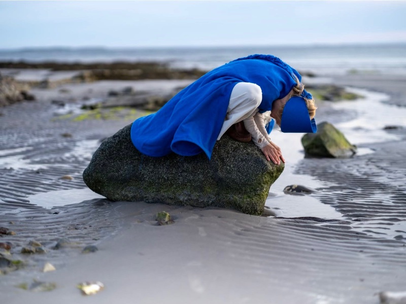 A woman dressed in blue kneeling on a big stone on the beach