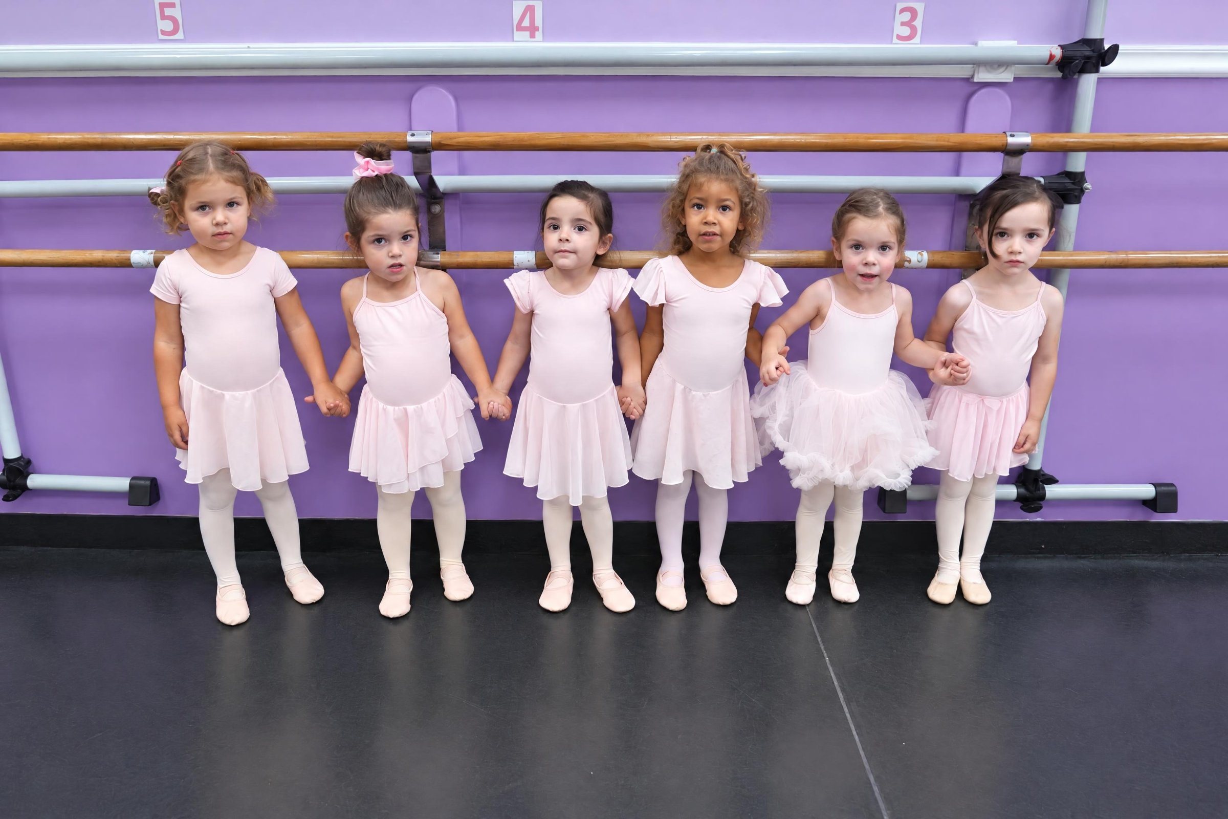 Six preschool ballet dancers in pink holding hands at the barre