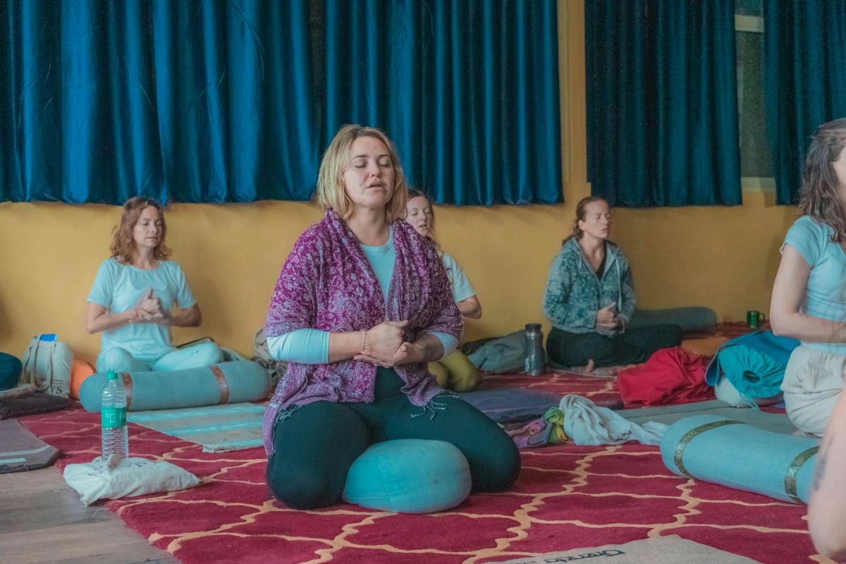 Woman meditating cross-legged among others in a room with blue curtains