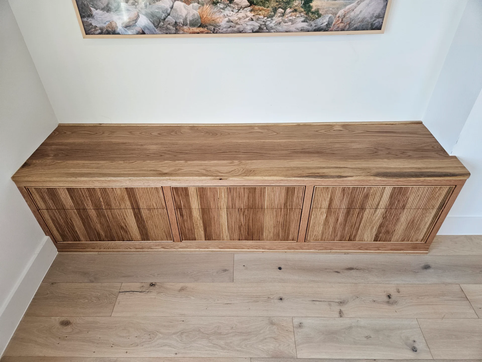 A custom built-in white oak cabinet with modern slatted door panels is set against a white wall in an Austin home. Above the credenza hangs a large mountain landscape painting, complemented by light-toned wood flooring in this West Lake Hills residence. - Detail 3