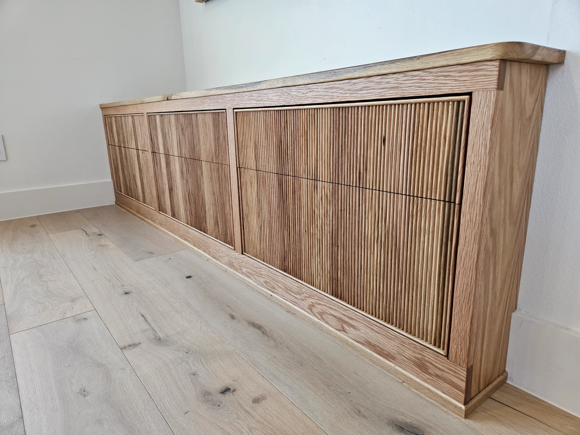 A custom built-in white oak cabinet with modern slatted door panels is set against a white wall in an Austin home. Above the credenza hangs a large mountain landscape painting, complemented by light-toned wood flooring in this West Lake Hills residence. - Detail 4
