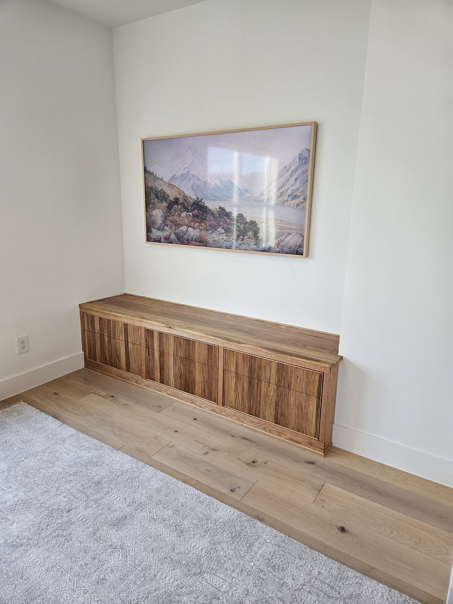 A custom built-in white oak cabinet with modern slatted door panels is set against a white wall in an Austin home. Above the credenza hangs a large mountain landscape painting, complemented by light-toned wood flooring in this West Lake Hills residence. - Detail 1