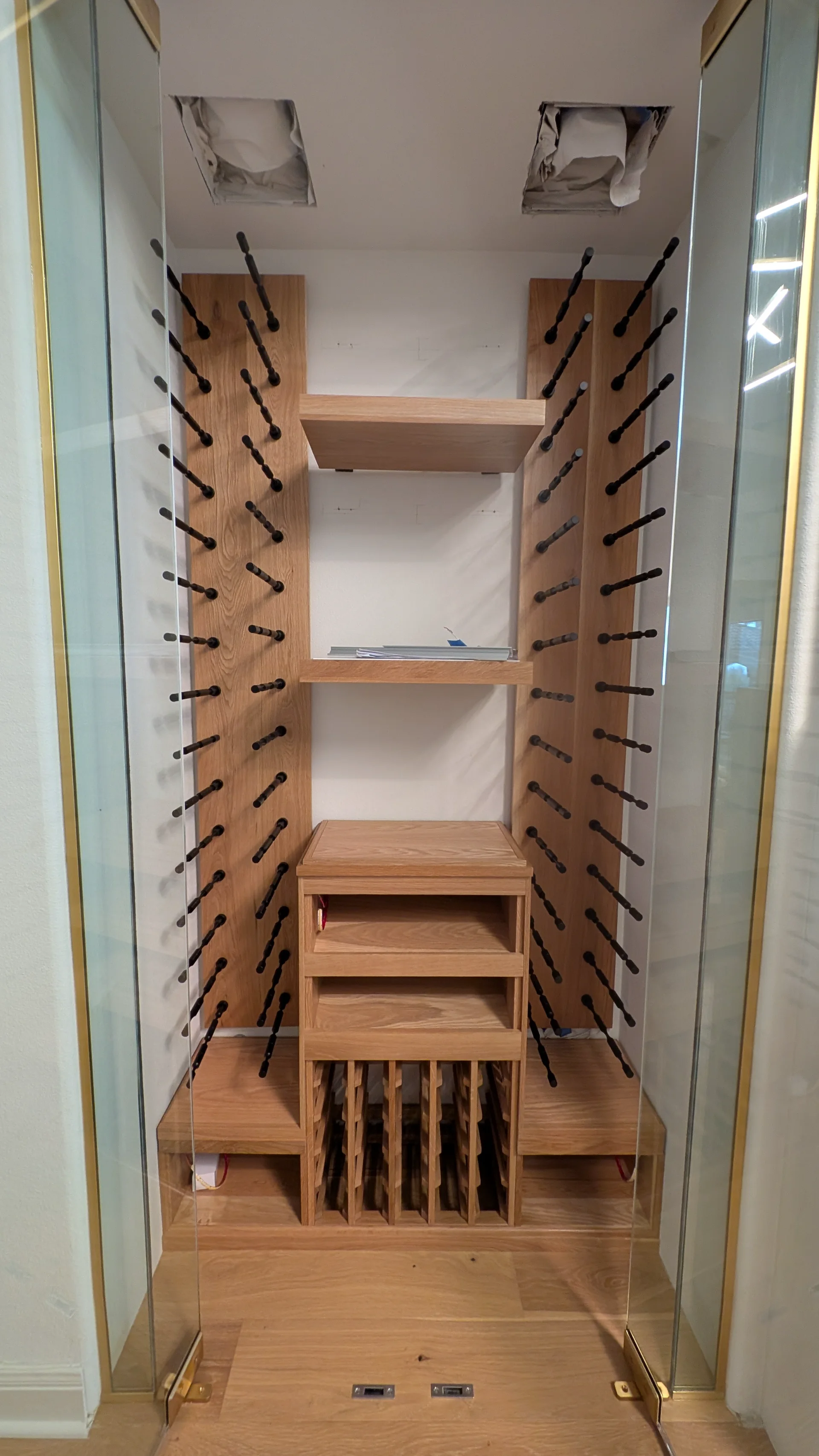A high-end custom white oak wine cellar under construction featuring modern black metal wine pegs and glass-enclosed shelving for a luxury home project in West Lake Hills, Austin. - Detail 1
