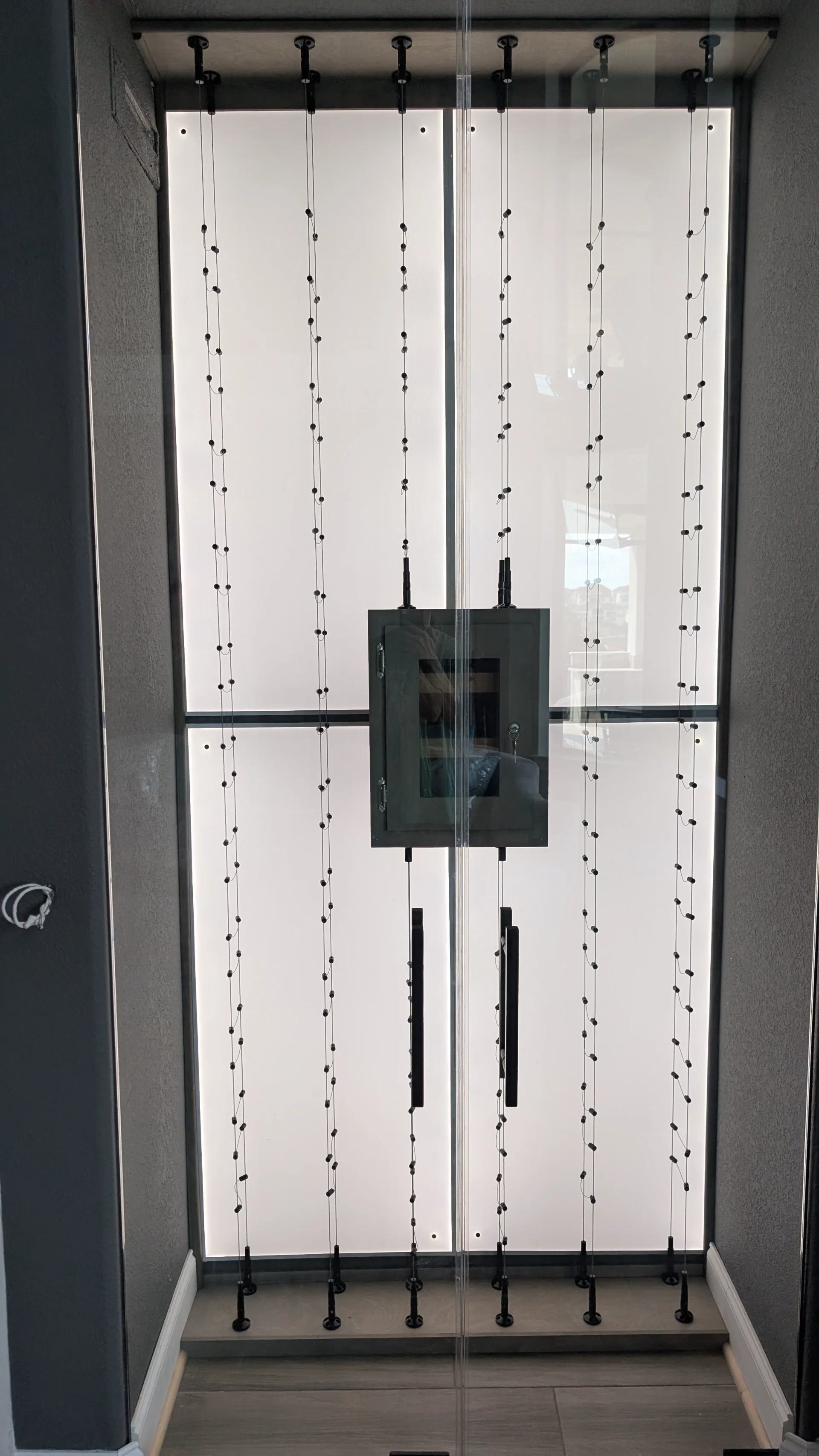 A high-end, contemporary glass wine cellar with a glowing, backlit frosted panel and a vertical cable racking system. This sleek architectural feature is part of a custom home project in Austin, West Lake Hills, and the Texas Hill Country. - Detail 1