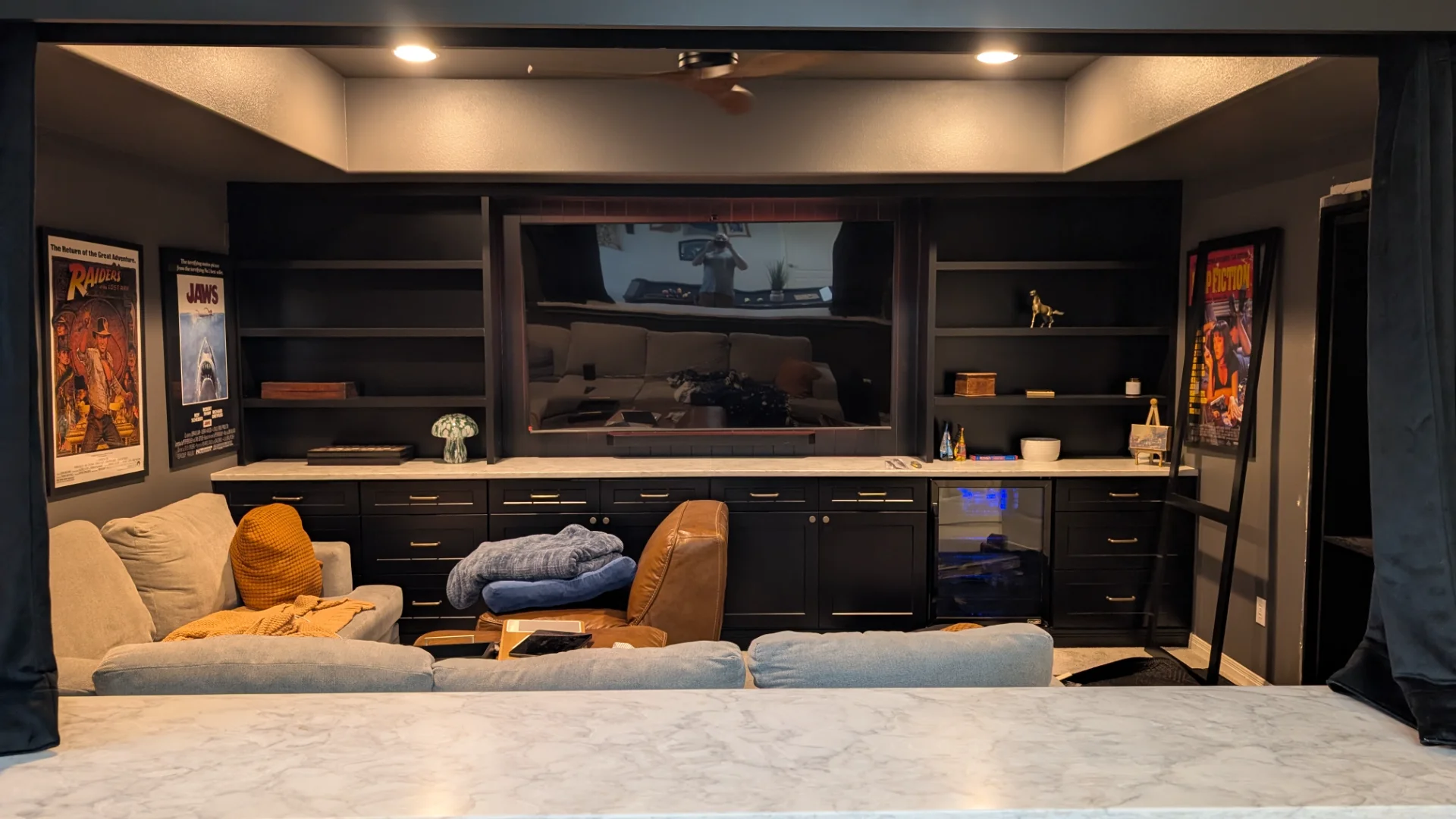 Sophisticated home theater in West Lake Hills, Austin, featuring a large custom black built-in entertainment center with marble countertops, an integrated wine fridge, and classic movie posters for Jaws and Pulp Fiction. - Detail 1