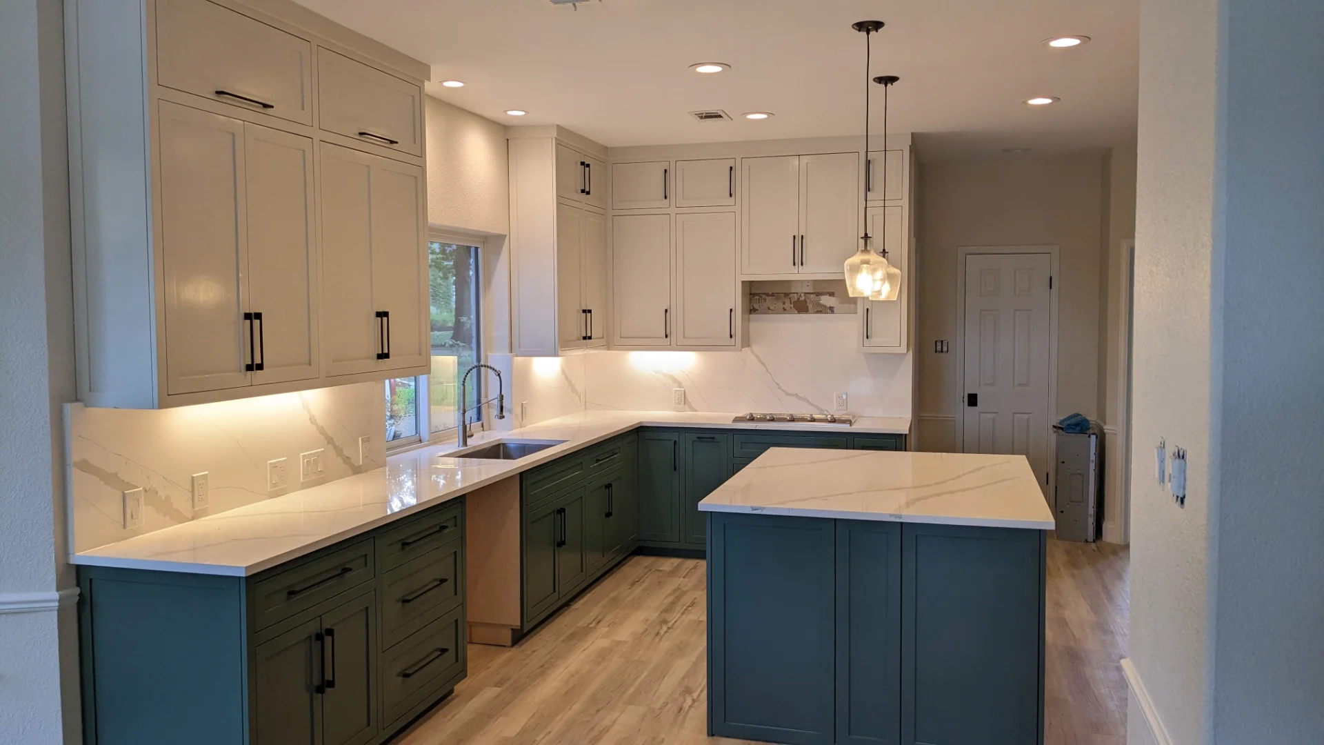 A modern two-tone kitchen remodel in Austin, Texas, showcasing off-white upper cabinets and deep teal lower cabinets with black hardware. The design features marble-patterned quartz countertops and backsplash, a central island, and recessed lighting in a Tarrytown home. - Detail 1