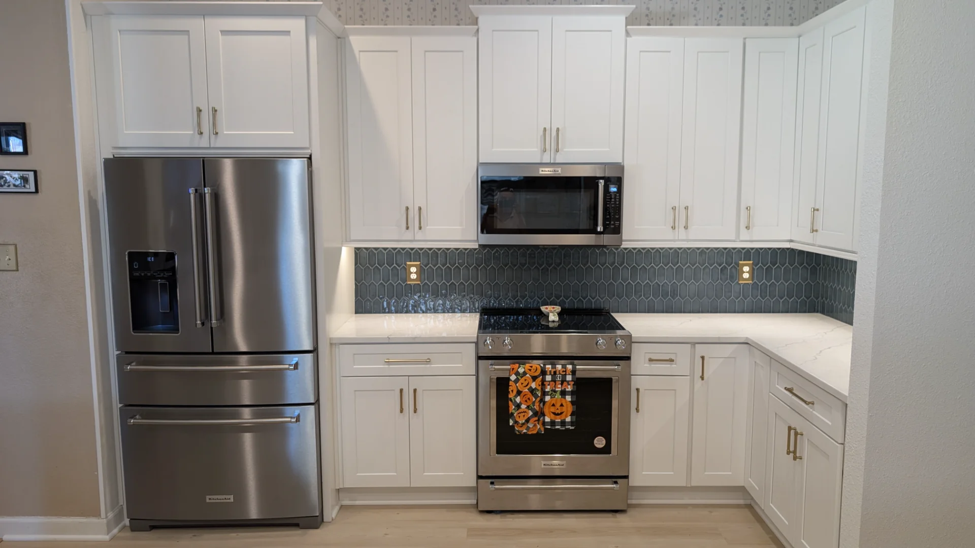 A modern kitchen remodel in Austin, Texas, featuring white shaker-style cabinetry with gold hardware, a blue-grey tile backsplash, white quartz countertops, and stainless steel KitchenAid appliances including a French door refrigerator and a slide-in range. - Detail 1
