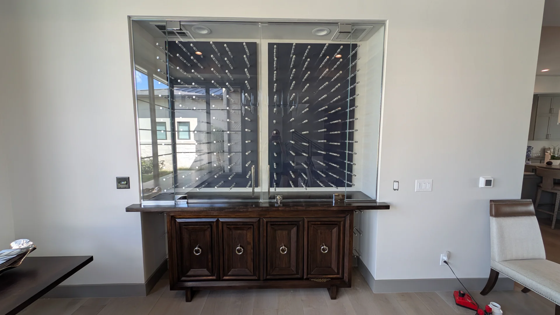 A modern, glass-enclosed wine cellar display with sleek chrome pegs against a dark navy background, set above a dark wood buffet cabinet in a luxury Austin, Texas home. - Detail 1