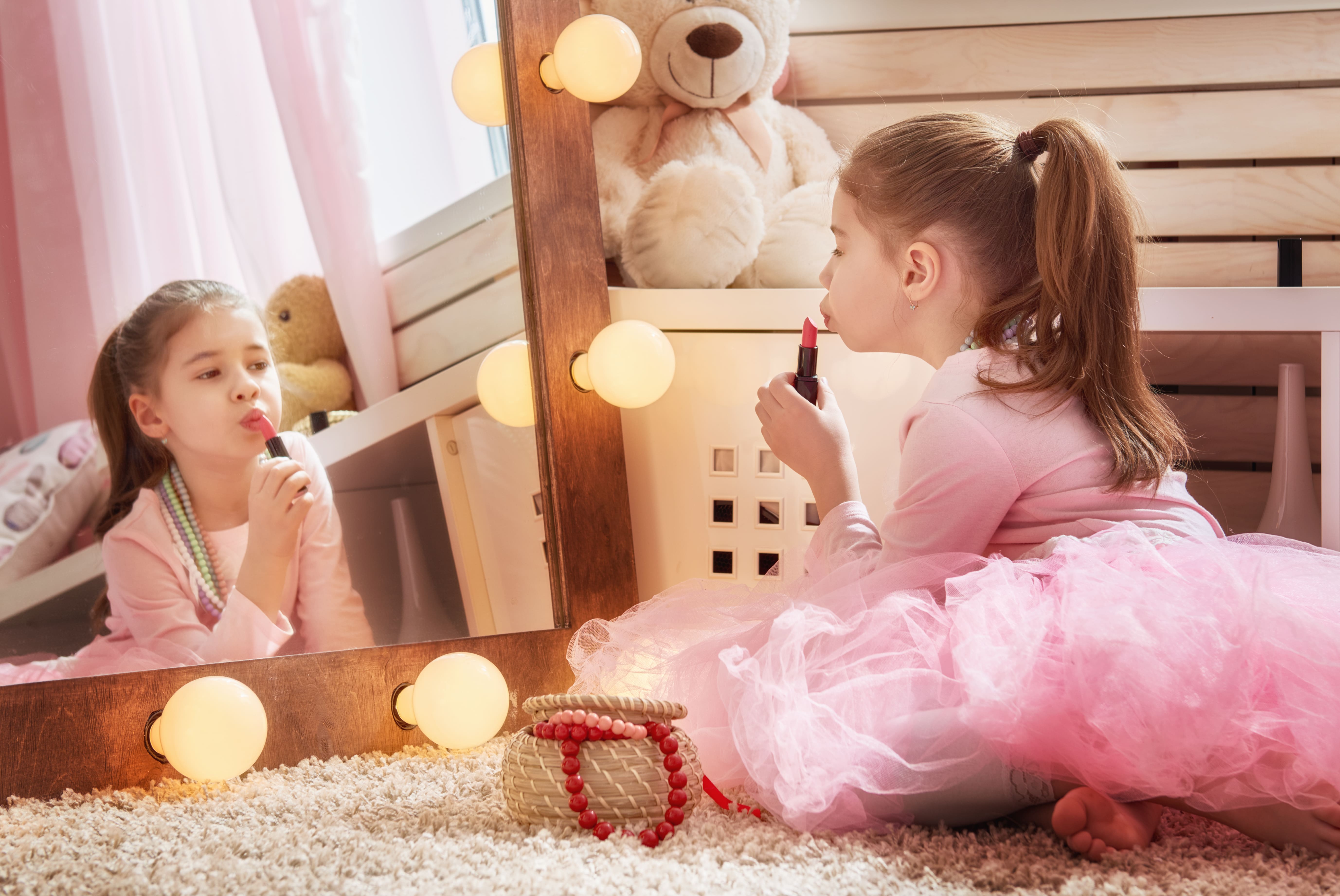 Young girl in a pink tutu with beaded necklace on, sitting on the floor, looking in the mirror while she puts on lipstick
