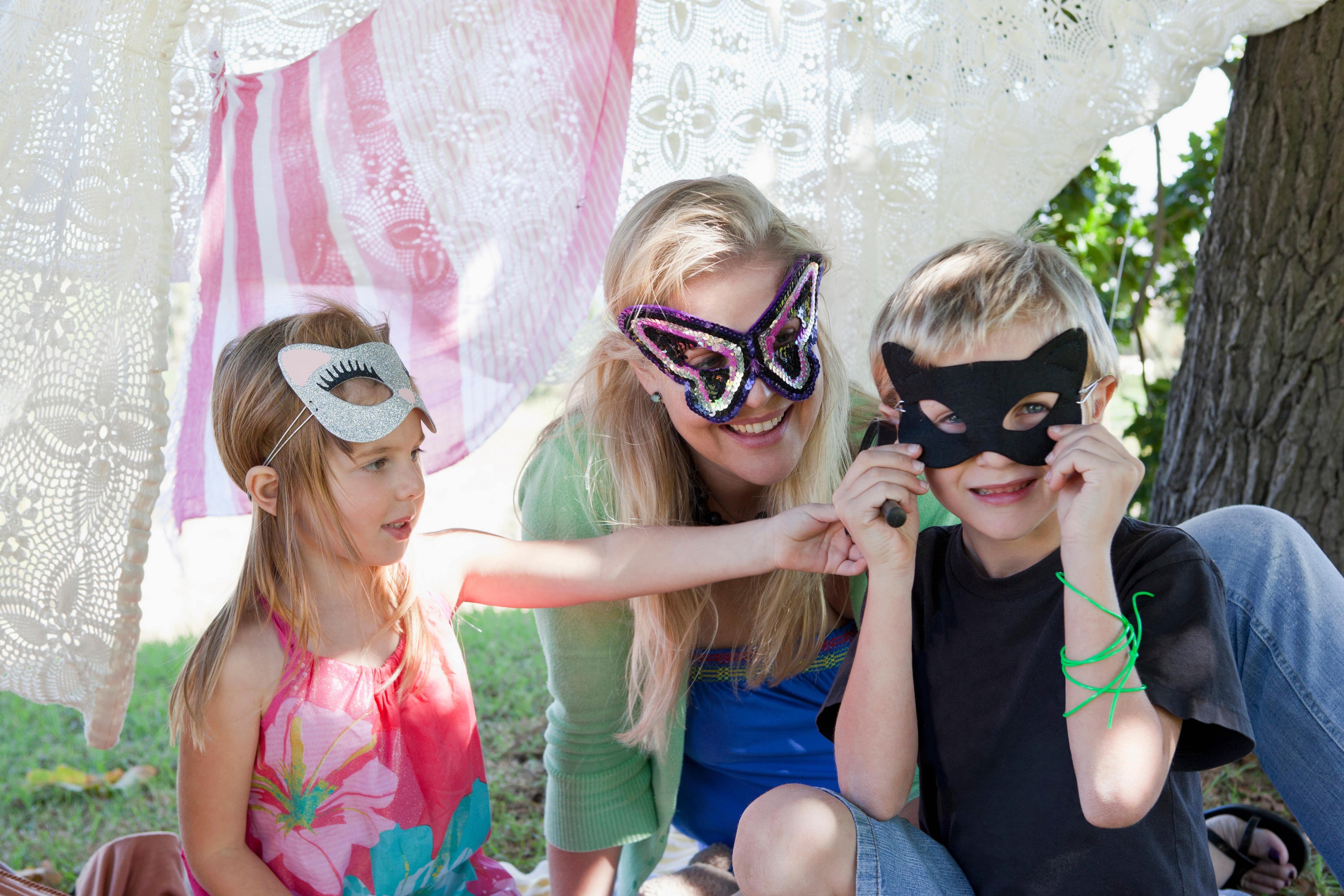 Young brother and sister sitting outdoors with mother, wearing birthday face masks
