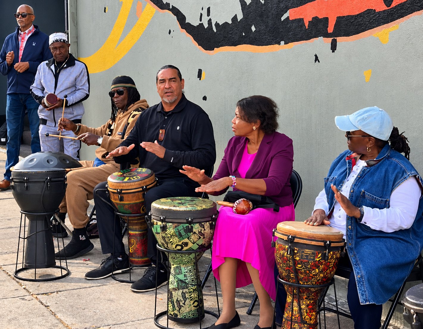 Members playing djembe drums outside beneath the mural