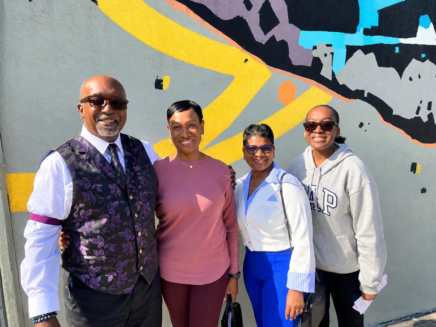 Four members smiling together in front of the Guidance mural