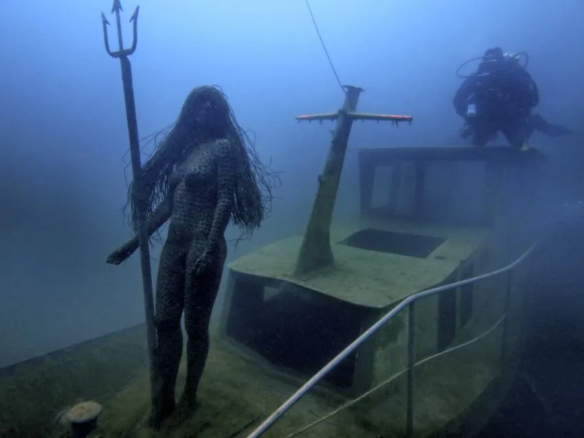 Statue mit Dreizack auf einem versunkenen Boot, dahinter ein Taucher im trüben Wasser.