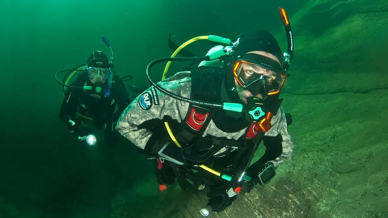 Zwei Taucher im Trockentauchanzug mit Lampen im grünen Wasser eines Sees.