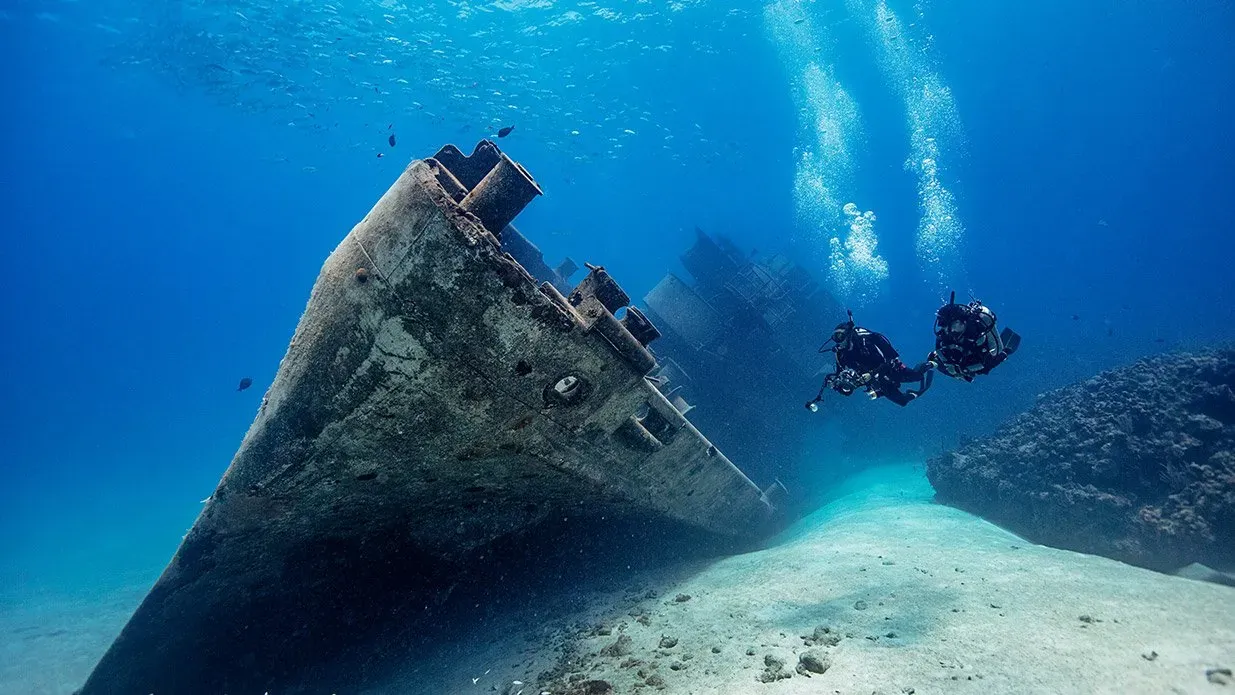 Zwei Taucher schweben über dem Bug eines großen Schiffswracks im klaren blauen Wasser.