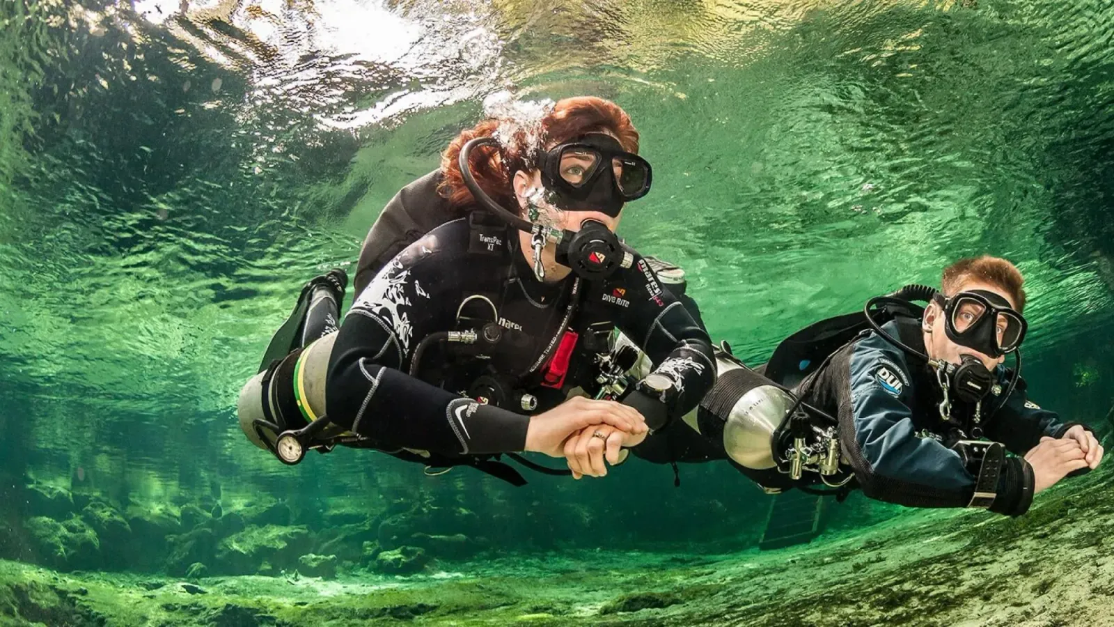 Zwei Taucher mit seitlich getragenen Flaschen gleiten dicht unter der Oberfläche durch klares grünes Wasser.
