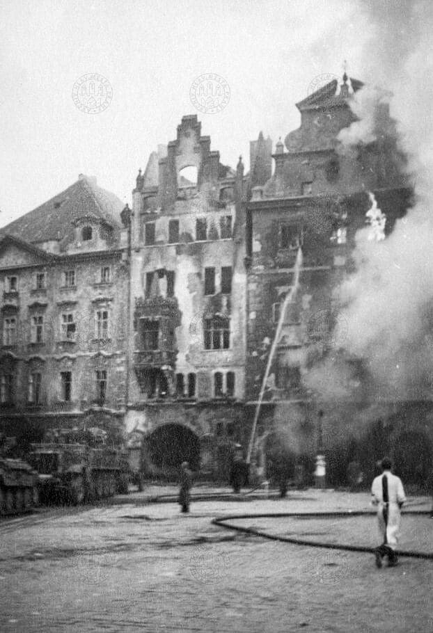 Жители Праги пытаются спасти горящие дома: Štorchův dům № 552 и dům U Kamenného beránka № 551 на Староместской площади. Тем временем мимо проходит колонна немецких солдат, покидающих Прагу 8 мая 1945 года.