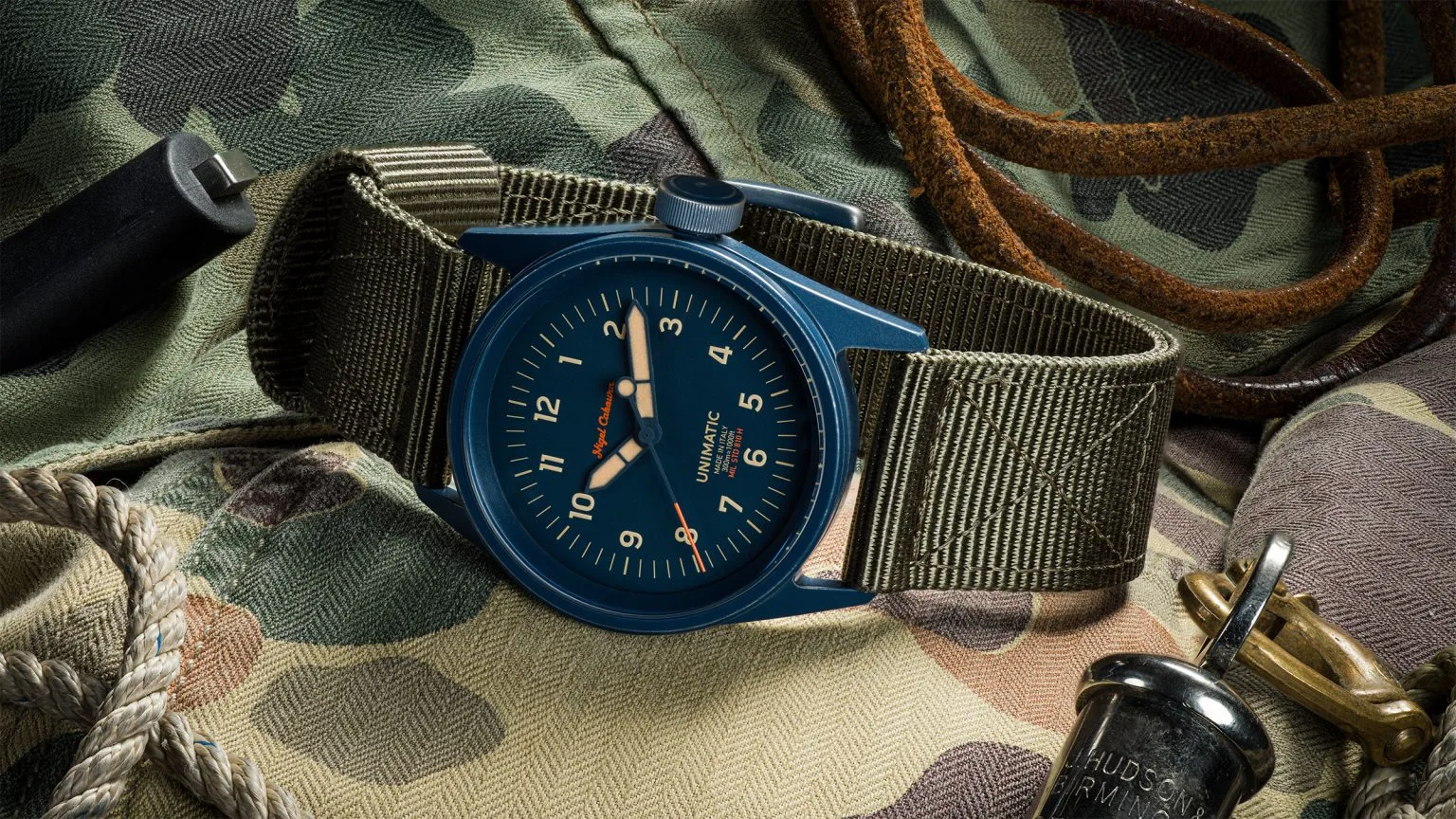 Blue Unimatic x Nigel Cabourn watch, green NATO strap, on camo fabric with rope, leather strap, and military accessories.