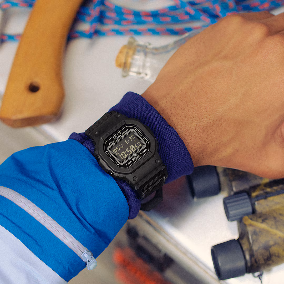 Close-up of a wrist wearing a black Casio G-Shock digital watch, with an axe handle, rope, bottle, and binoculars.
