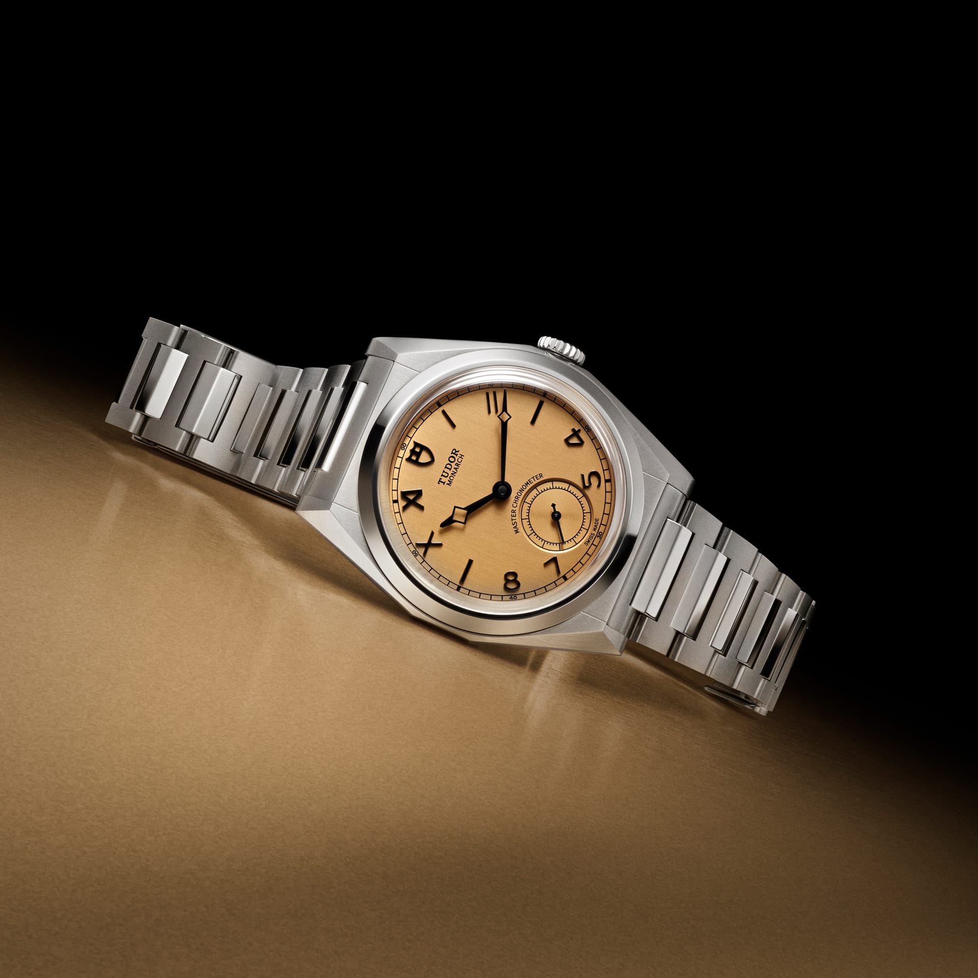 Angular stainless steel Tudor Monarch watch, golden dial with numerals and sub-seconds, on a reflective bronze surface.