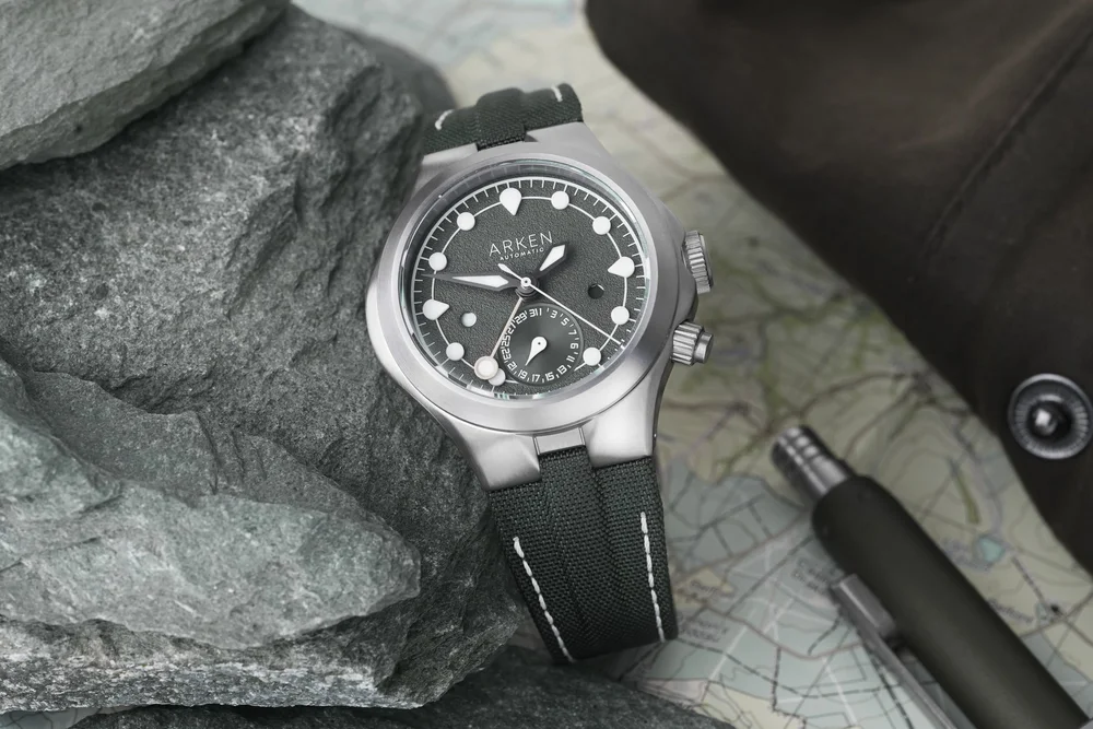 Arken Automatic steel watch with dark grey dial, white indices, green canvas strap, on rocks, map, pen, & bag.