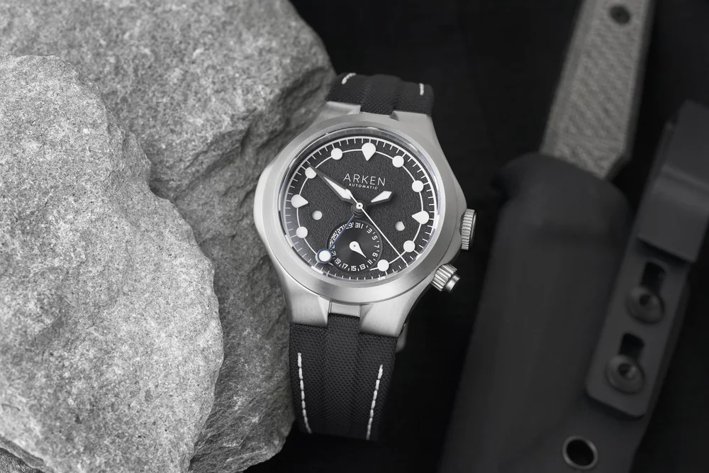 Arken Automatic watch with black dial, silver case, black strap, displayed on grey rocks with a knife & sheath.