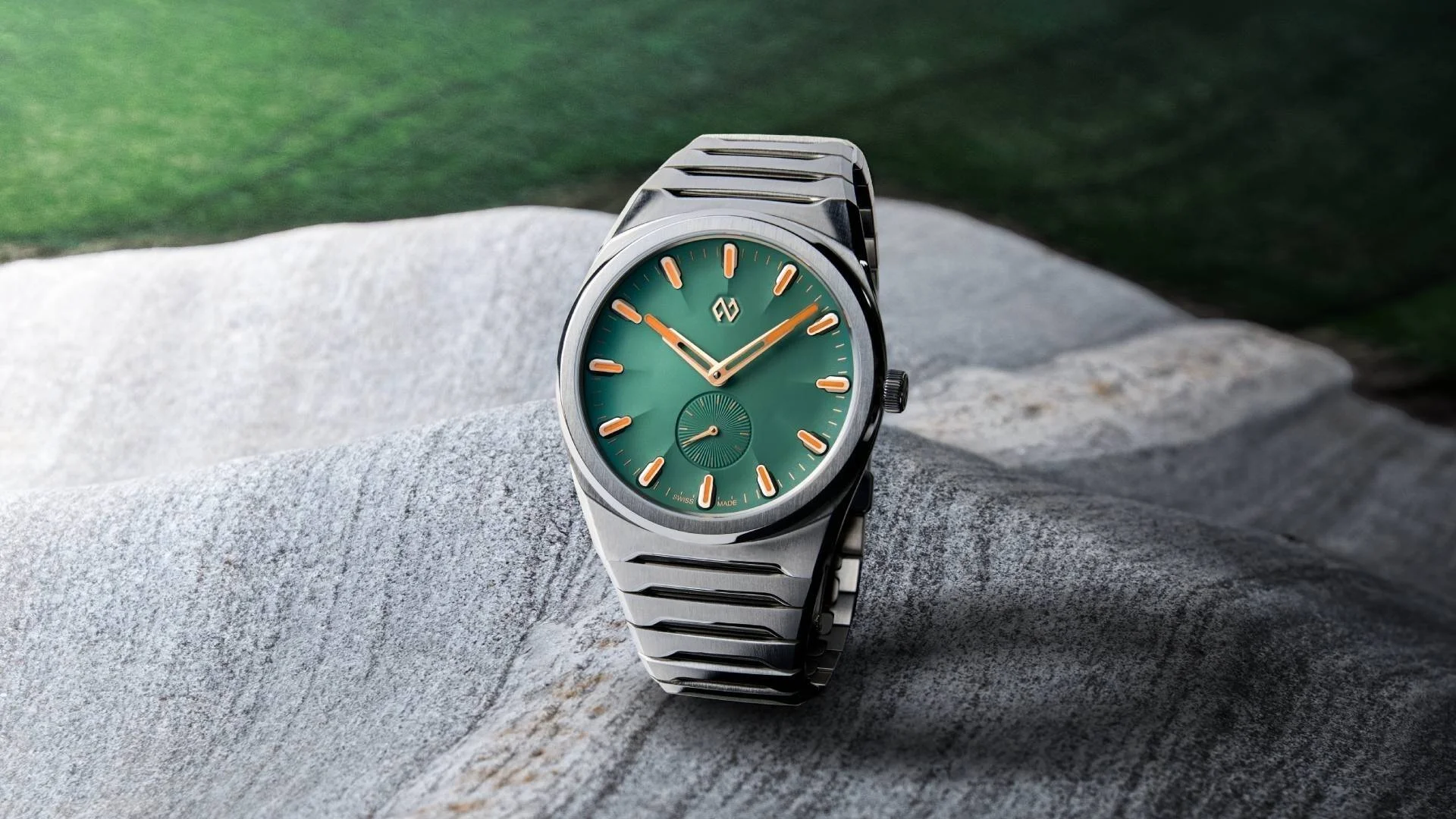Stainless steel luxury watch with green dial, orange hands & markers, and integrated bracelet on grey rocks with green background.