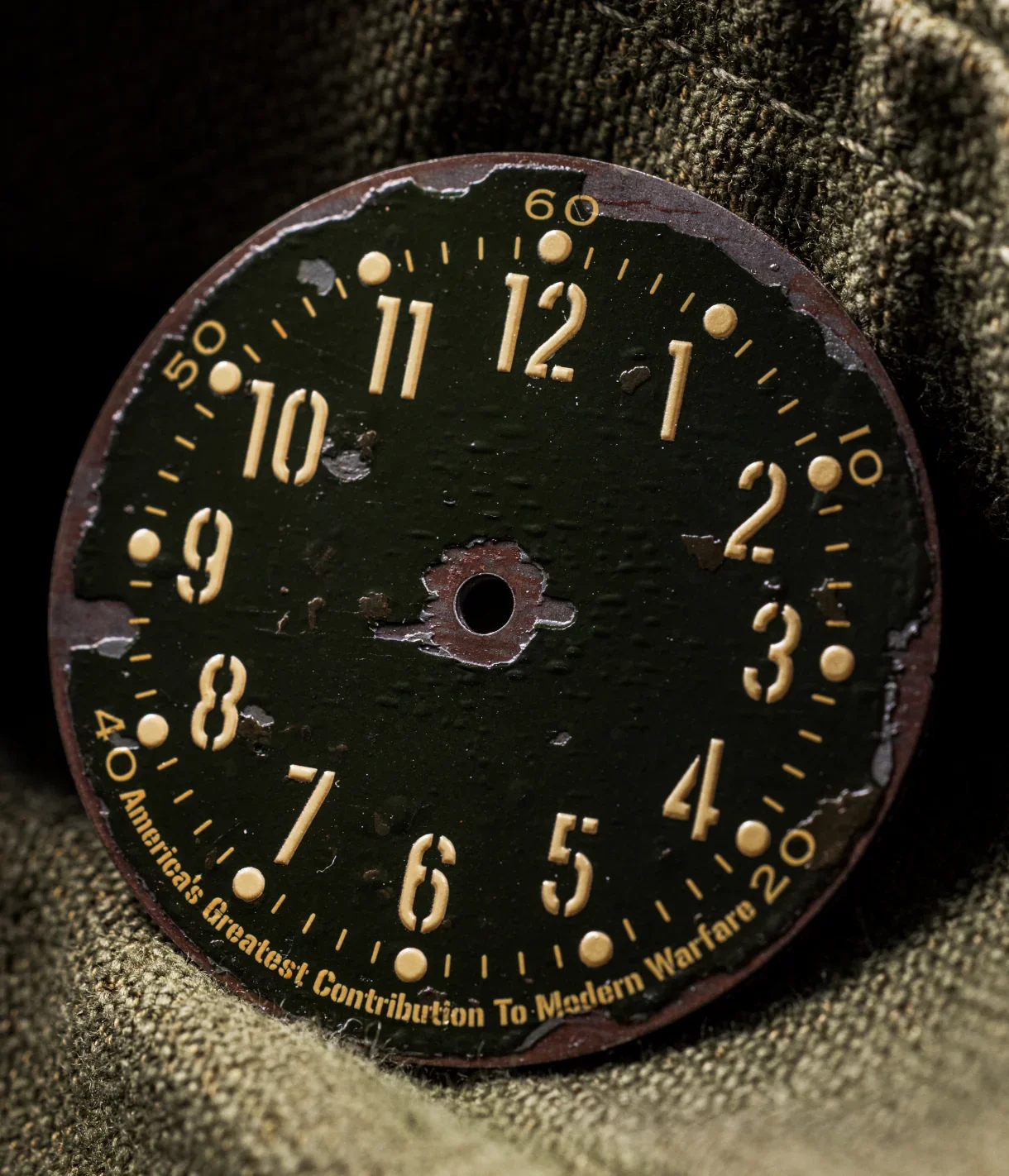 Worn olive green watch dial with yellow Arabic numerals, 'America's Greatest Contribution To Modern Warfare' text, on olive fabric.
