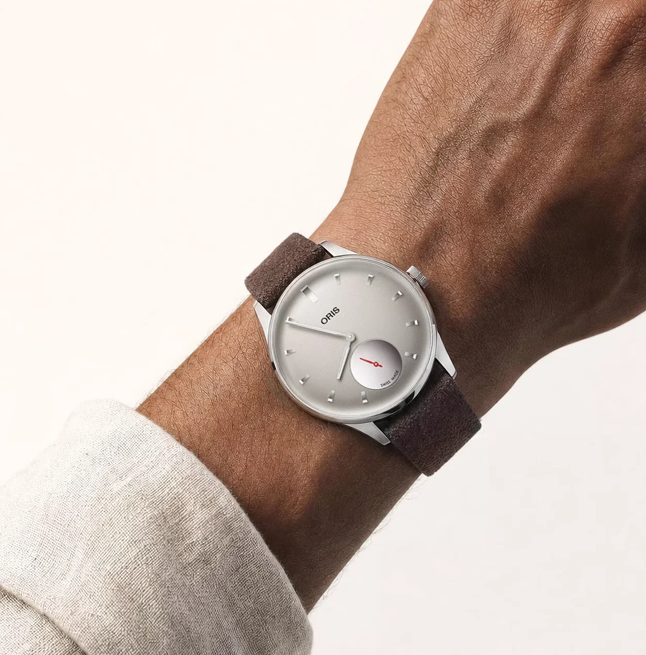 Oris watch on a person's arm: silver dial, brown strap, red sub-dial hand, light shirt cuff visible.