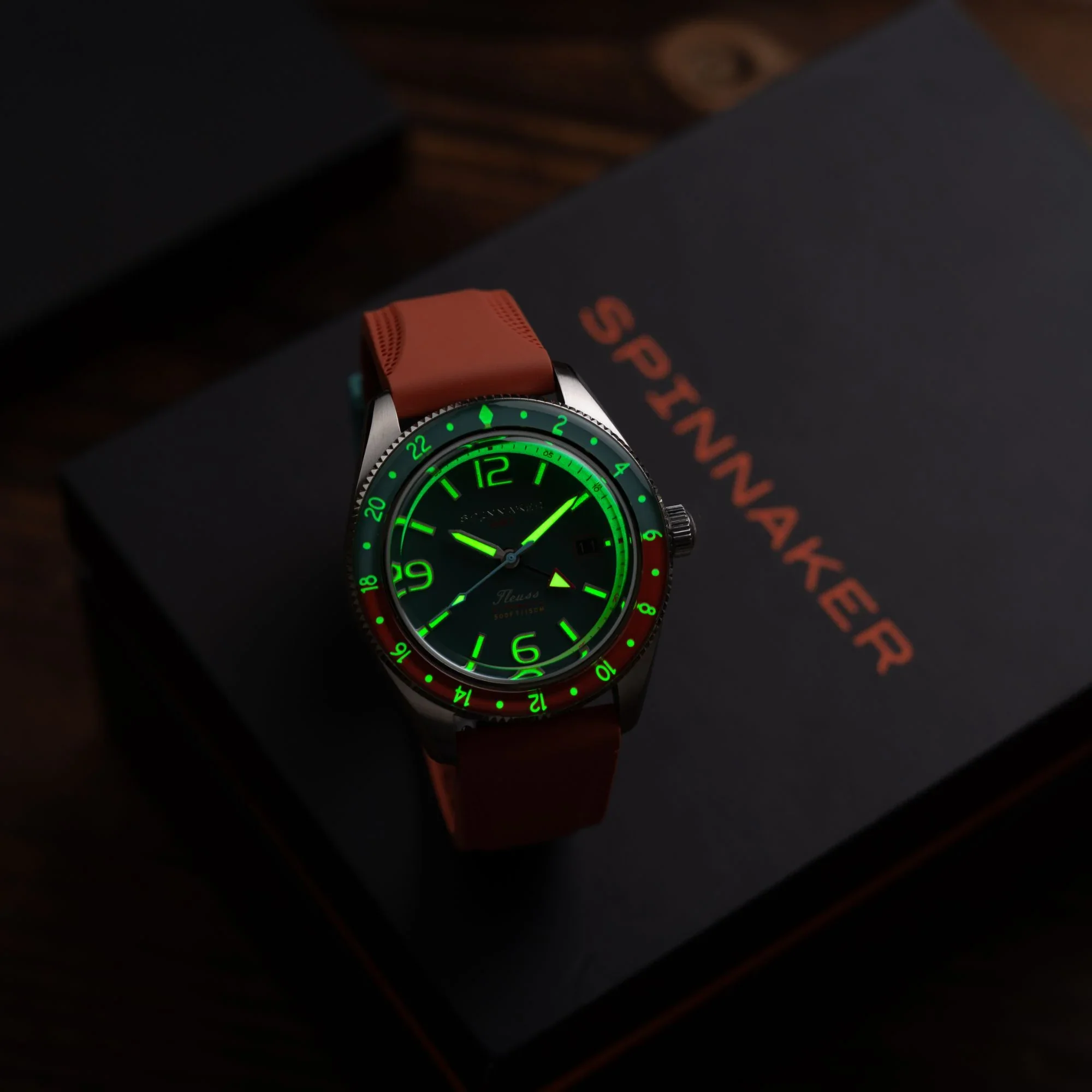 Spinnaker Fleuss GMT watch with glowing green dial, two-tone green/red bezel, and orange strap on a black Spinnaker box.