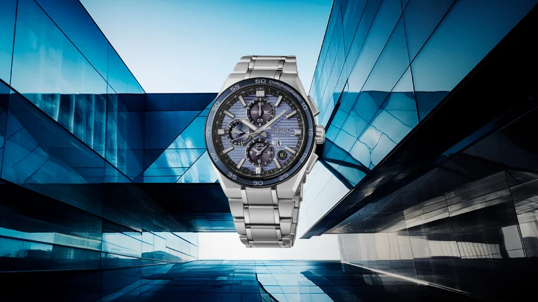 Seiko Astron chronograph watch with textured blue dial, silver-tone bracelet, set against a modern blue glass building.