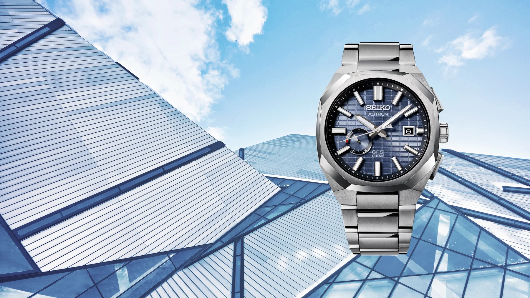 Seiko Astron GPS Solar watch, silver-toned with a blue textured dial, against modern angular buildings under a blue sky.