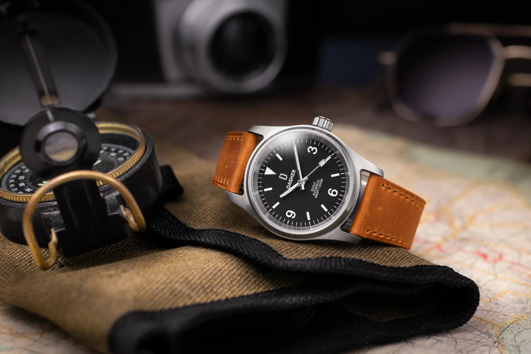 Silver Dasher Scout watch with black dial, brown leather strap on map. Compass, camera, sunglasses in background.