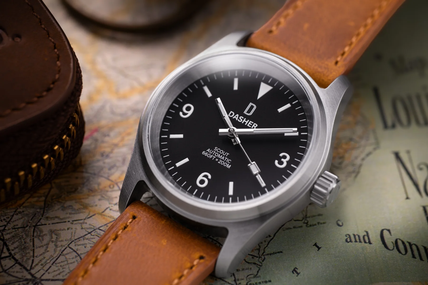 Dasher Scout 36mm automatic field watch with black dial & brown strap, on a map next to a zipped leather case.