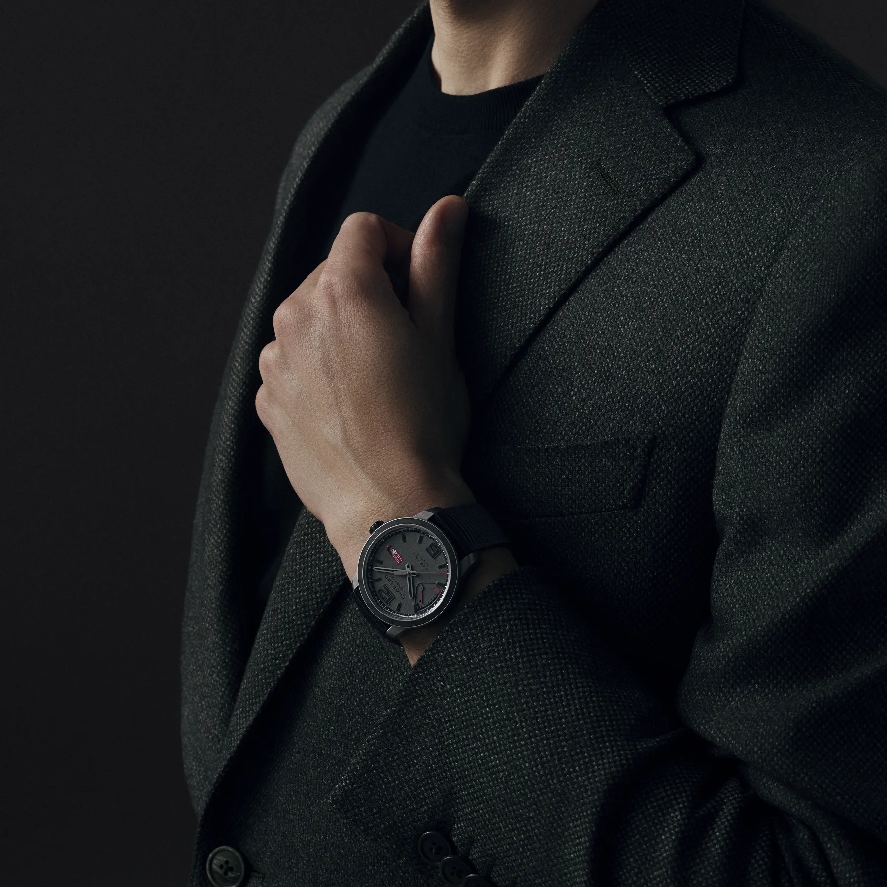 Man in textured dark grey blazer & black top, wearing a Chopard watch with grey dial, red accents, and black strap.