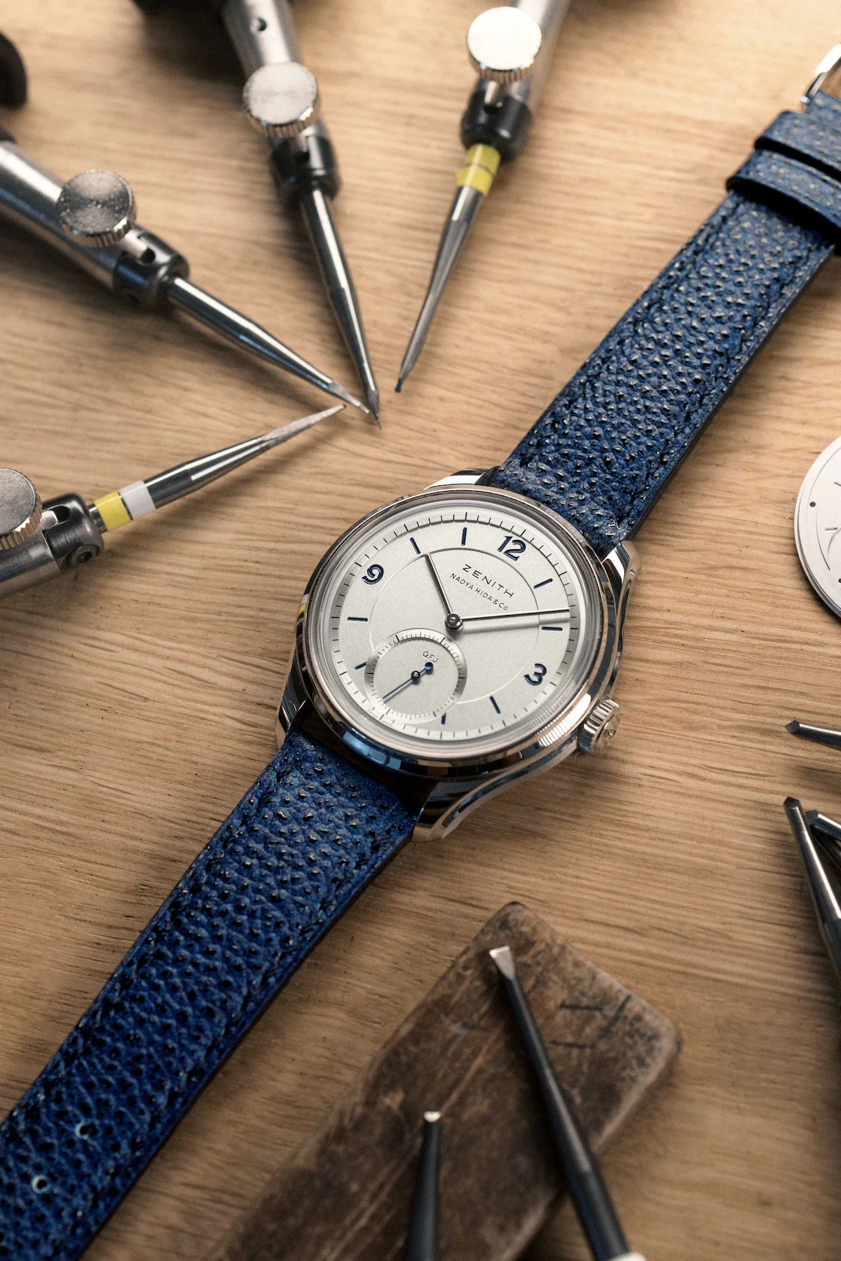 Zenith Naoya Hida & Co watch with white dial, blue textured strap, & watchmaking tools on wooden bench.