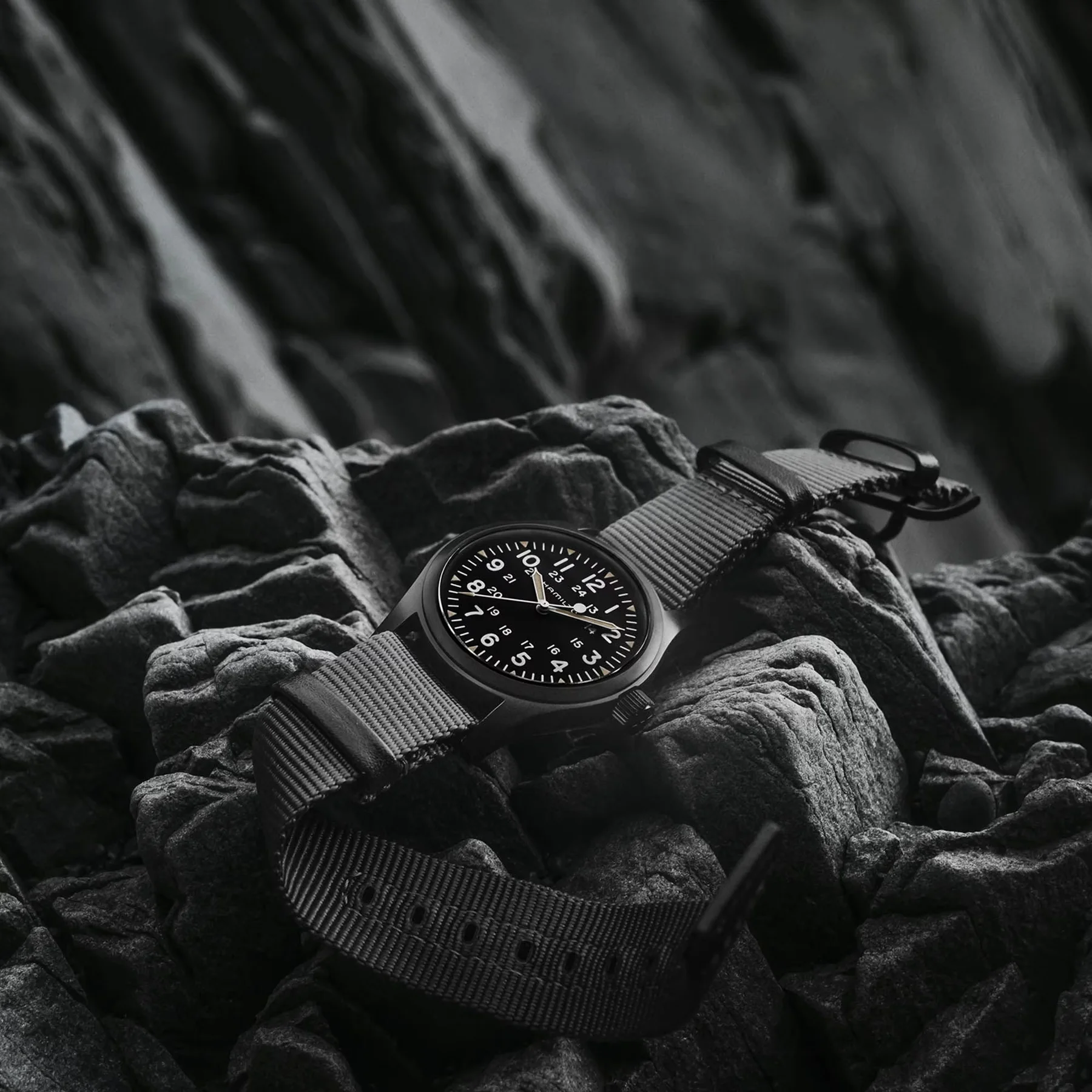 Black Hamilton field watch with white numerals & a dark fabric strap on textured grayscale rocks.