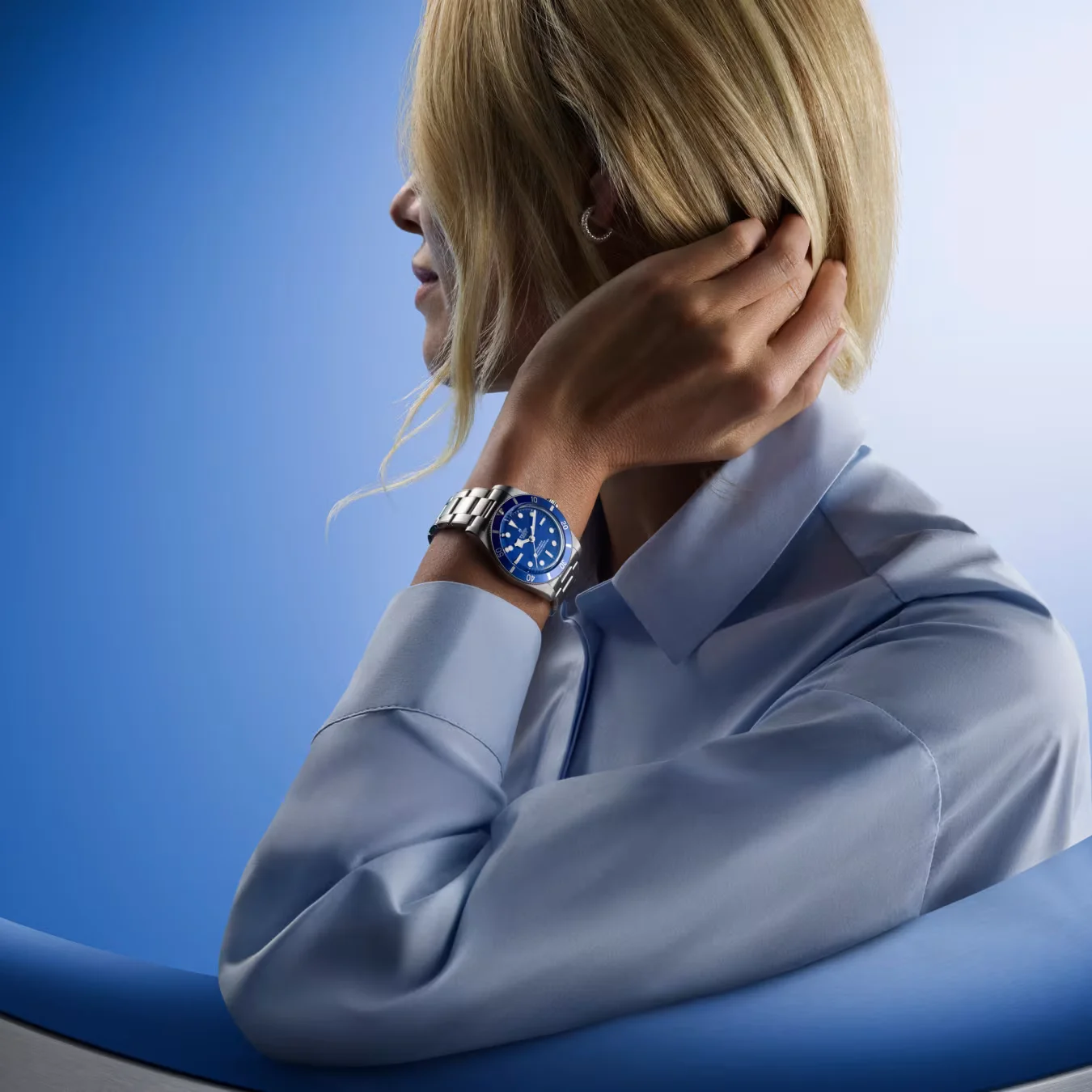 Blonde woman in blue shirt wearing a blue Tudor Black Bay 54 on a steel bracelet, against a gradient blue background.