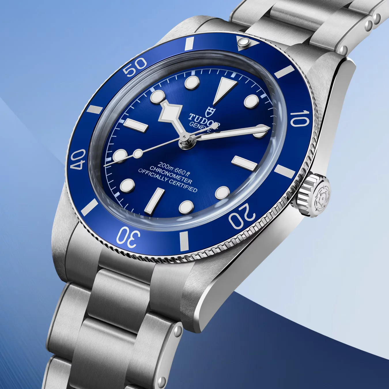 Arm of person in blue denim shirt & white pants, wearing blue-dial Tudor Black Bay 54 on steel bracelet. Blue background.