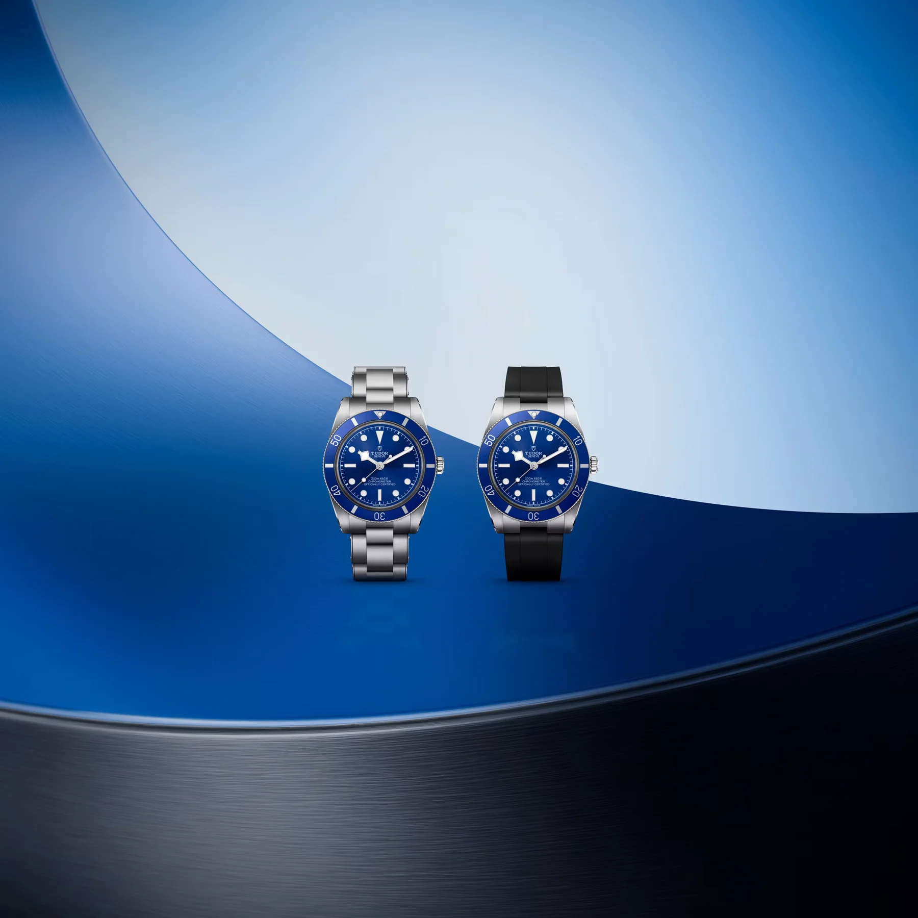 Two blue Tudor Black Bay 54 watches: one on a steel bracelet, one on a black rubber strap, on blue background.