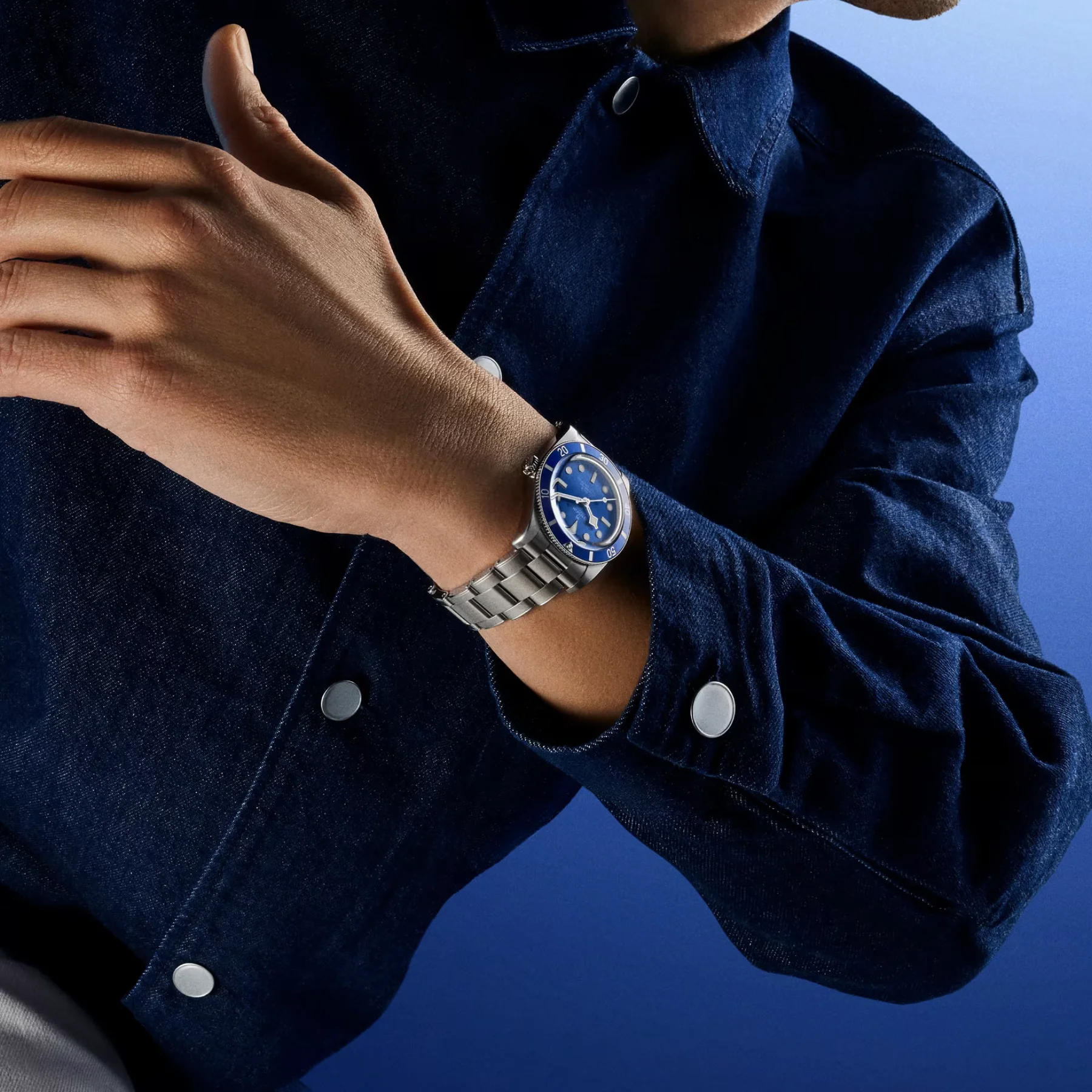 Arm in denim jacket wears blue Tudor Black Bay 54 on steel bracelet against blue background.