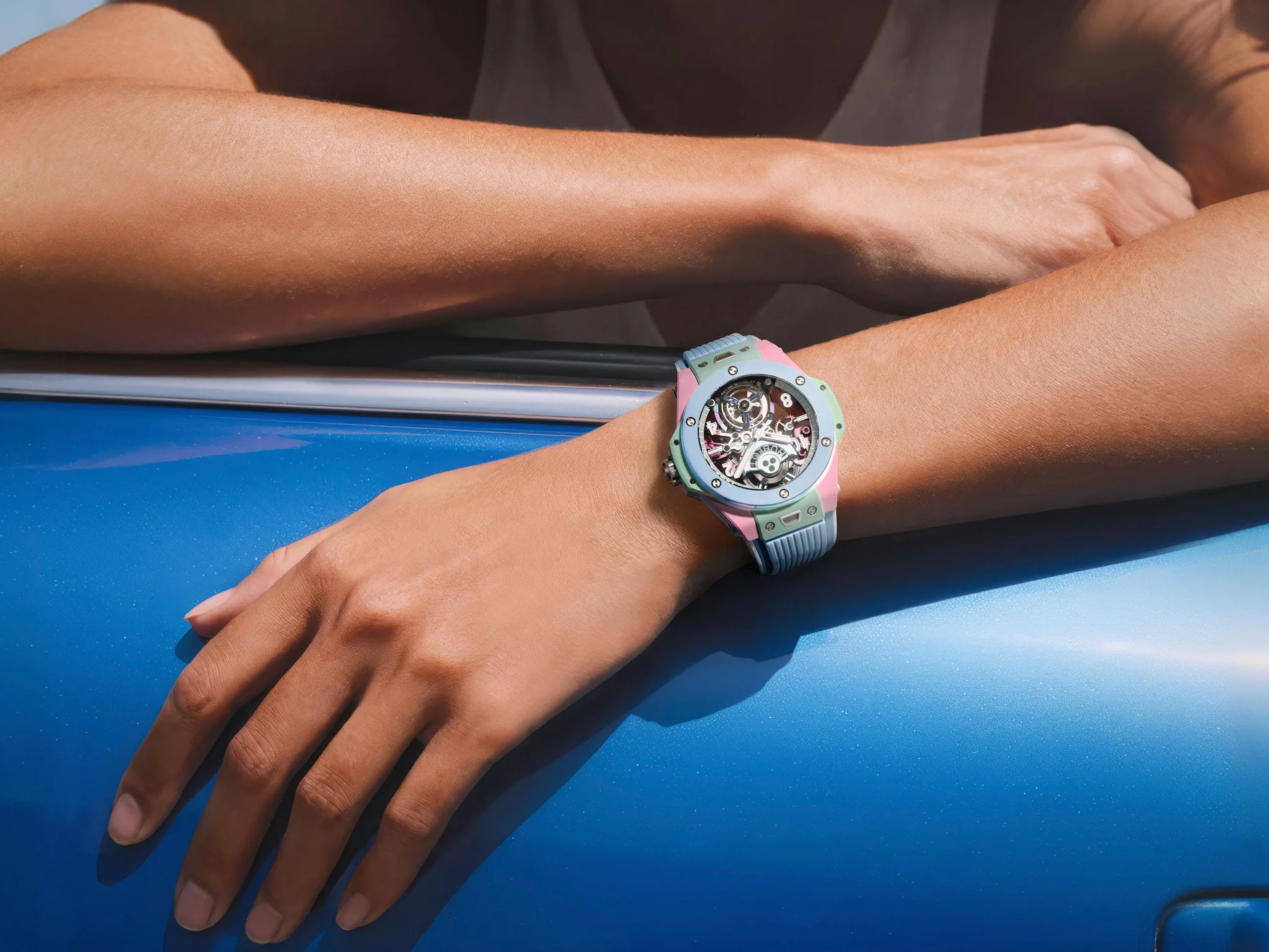 Close-up of an arm wearing a pastel Hublot Big Bang Unico skeleton watch, leaning on a shiny blue surface.